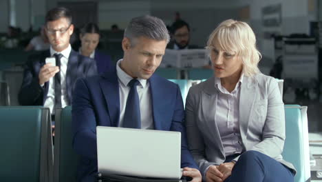 Primer-Plano-De-Dos-Colegas-Usando-Una-Computadora-Portátil-En-El-Vestíbulo-Del-Aeropuerto,-Otros-Pasajeros-Esperando-El-Vuelo-En-El-Fondo