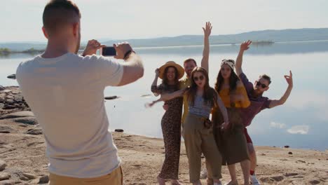 group of happy young people standing on sand by peaceful lake and posing for photograph together