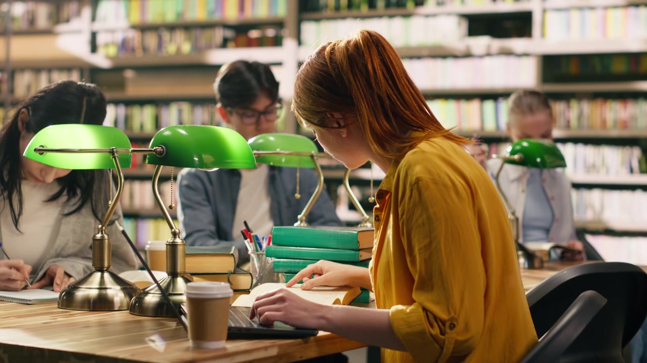 Studentessa dai capelli rossi che beve caffè mentre usa il portatile alla scrivania della biblioteca.