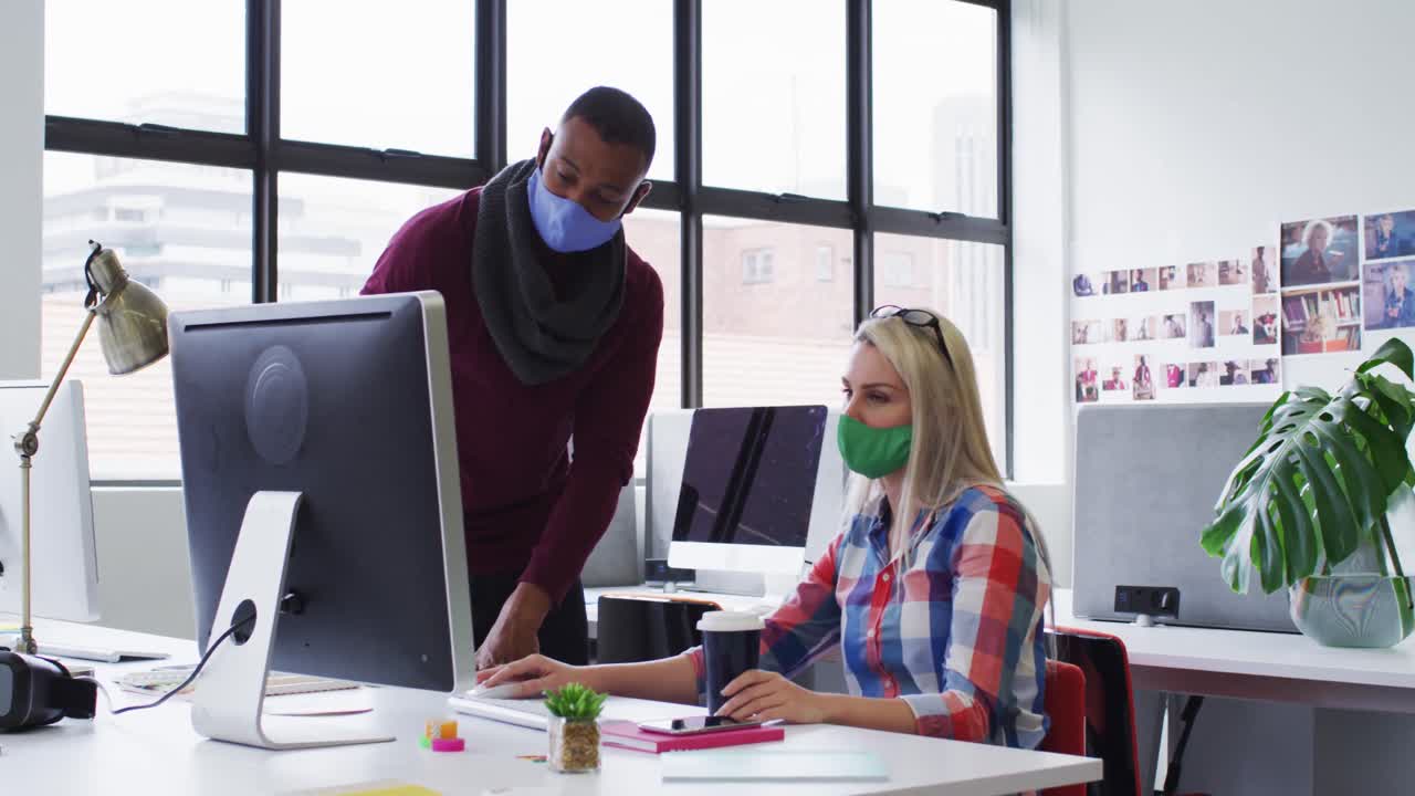 Diverse male and female office colleagues wearing face masks using computer at modern office