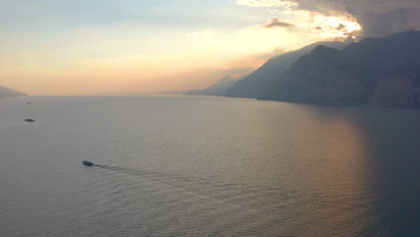 Statische,-Stimmungsvolle-Luftaufnahme-Eines-Großen-Bootes,-Das-Bei-Sonnenuntergang-über-Den-Gardasee-Segelt-Und-Der-Landschaft-Ein-Gefühl-Von-Weite-Verleiht