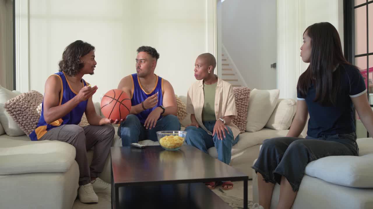 Diverse friends bringing chips on couch, reaching and tossing orange basketball while watching TV