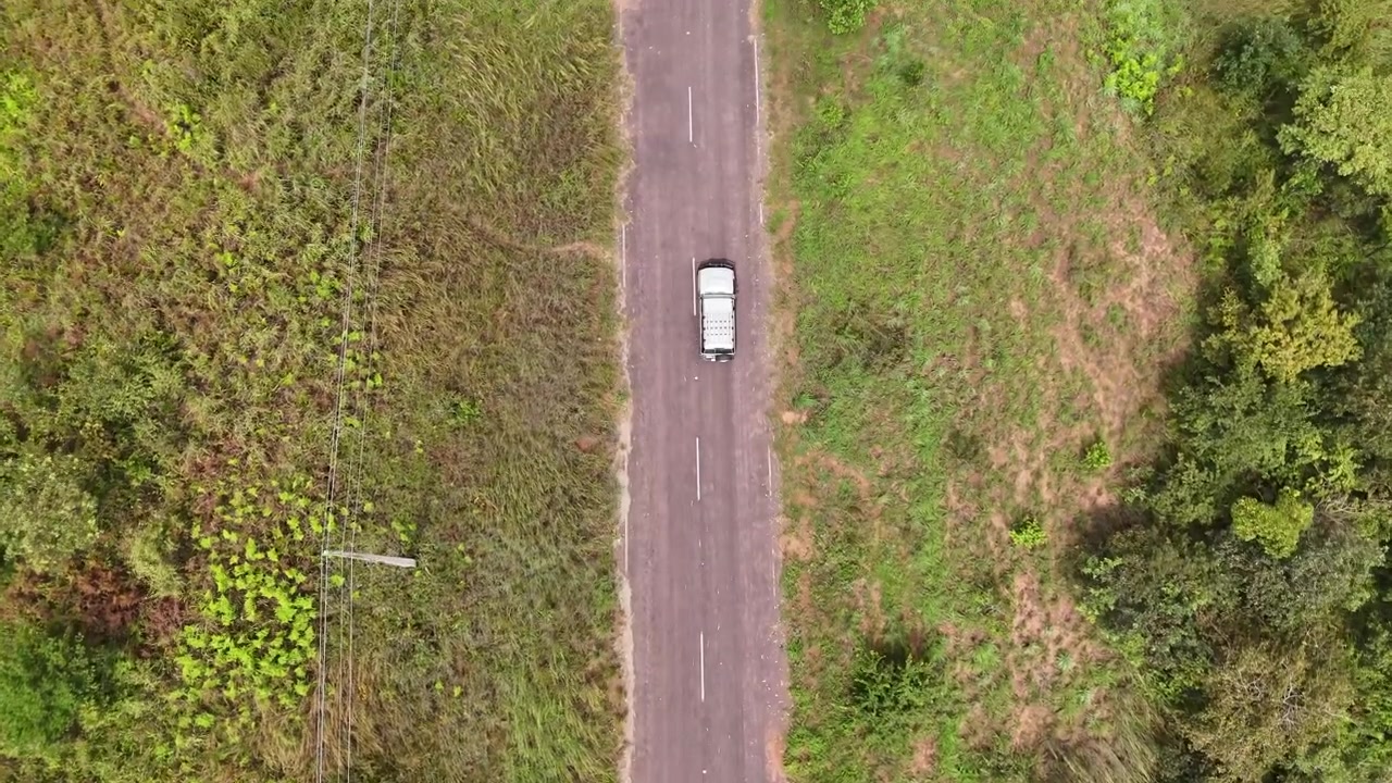 Ripresa aerea zenitale diretta di un pick-up bianco che percorre una strada asfaltata rurale diritta, attraversando rigogliosi campi erbosi verdi e vegetazione campestre in Angola, Africa.