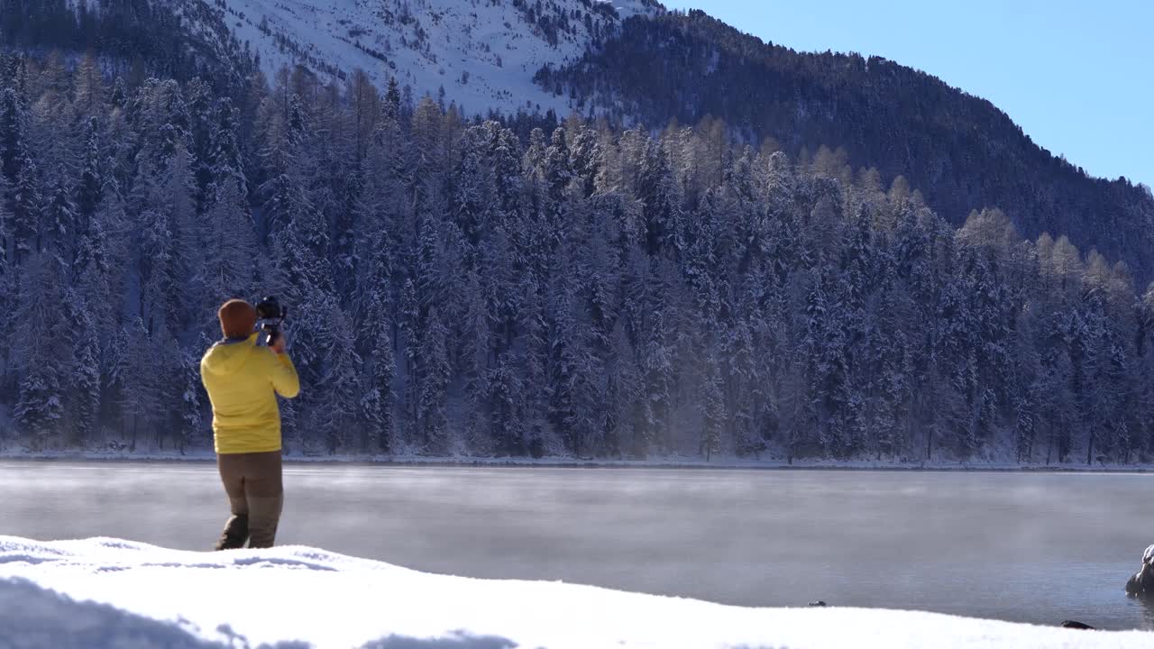 joven caucásico grabando un video con una cámara en un cardán junto al lago cubierto de nieve y árboles en st.