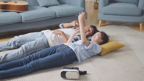 asian teen couple using smartphone taking selfie while lying on carpet on the floor at home. smiling, showing peace and heart gesture