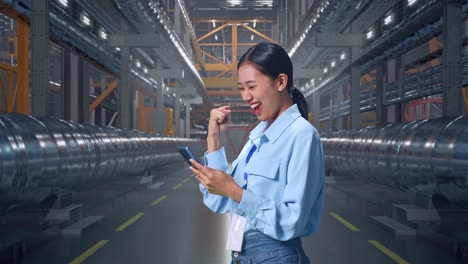 side view of an asian female professional worker with her smartphone in a in big warehouse filled with metal pipes, she raises her fist up with screaming goal after check on the smartphone