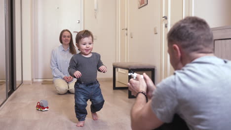 Madre-Y-Su-Bebé-En-Un-Extremo-Del-Pasillo-En-Casa-Mientras-El-Padre-Anima-Al-Bebé-A-Caminar-Hacia-El-Otro-Extremo-2