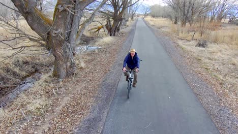 Mature-cyclist-biking-along-a-nature-trail-beside-a-creek-and-between-trees---leading-aerial-view