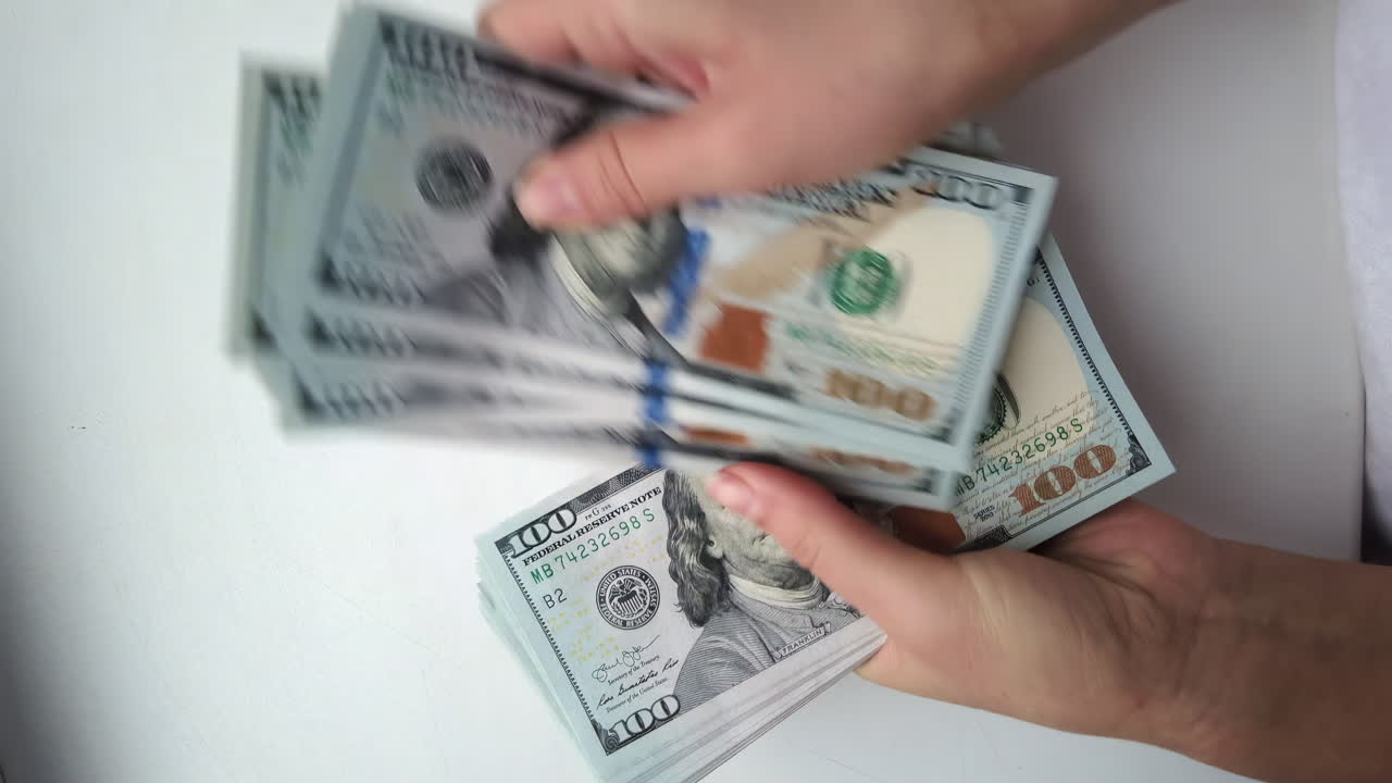 Hands organize and count stacks of hundred-dollar bills in a well-lit room. The focus is on the careful handling and arrangement of the cash, conveying a sense of precision
