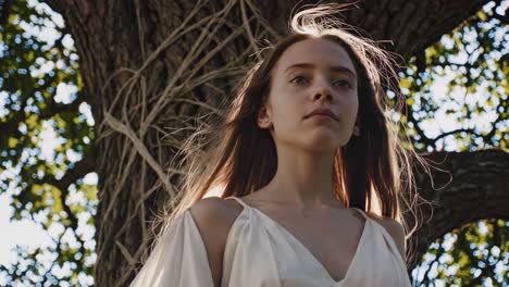young woman in a flowing white dress posing gracefully near a large tree, adorned with string, during a captivating theatrical performance in a sunlit forest, creating a magical atmosphere