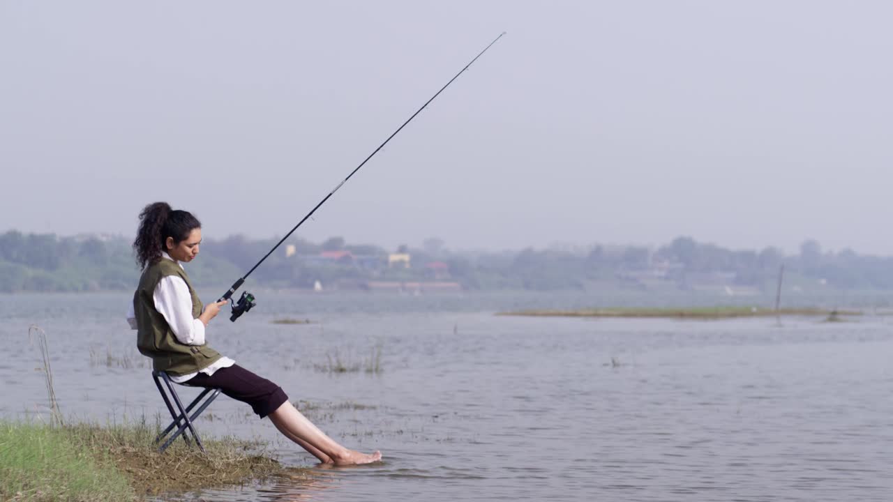 Seorang gadis Asia Selatan mengenakan rompi luar ruangan sedang duduk memancing di tepi danau yang tenang, bersantai dengan kakinya di air dangkal