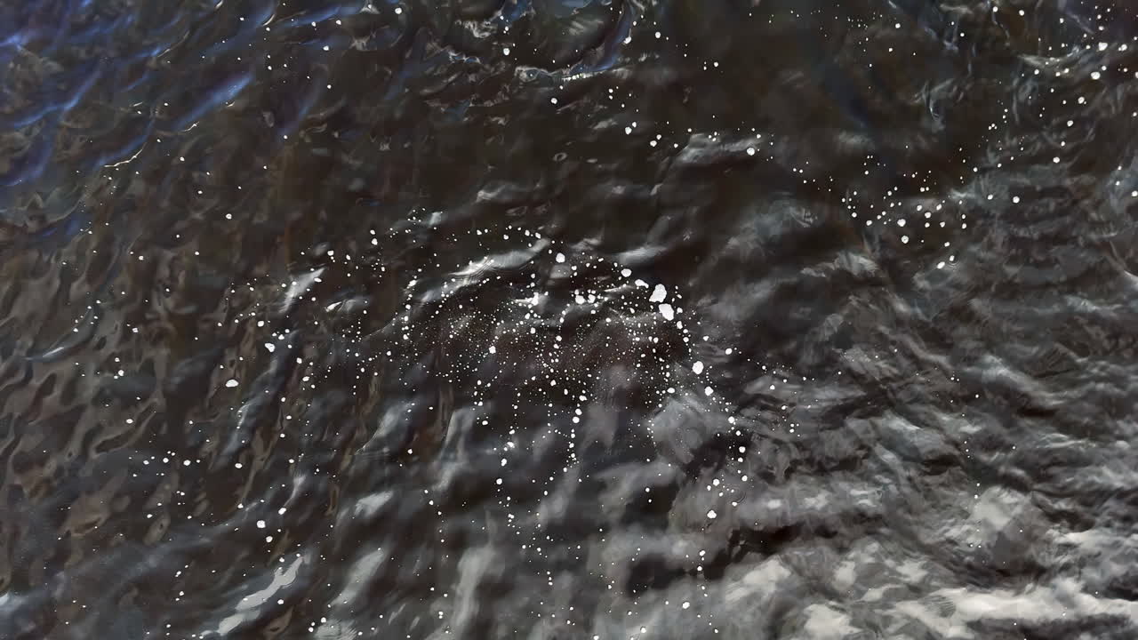 Aerial view of bubbling floodwaters flowing over dark terrain. Surface foam and ripples indicate turbulent movement, evoking the chaos and force of storm runoff or flash flooding