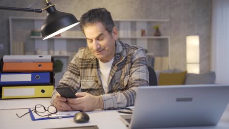 Smiling-mature-man-working-in-home-office-looking-at-phone,-texting,-surfing-social-media-and-being-happy.