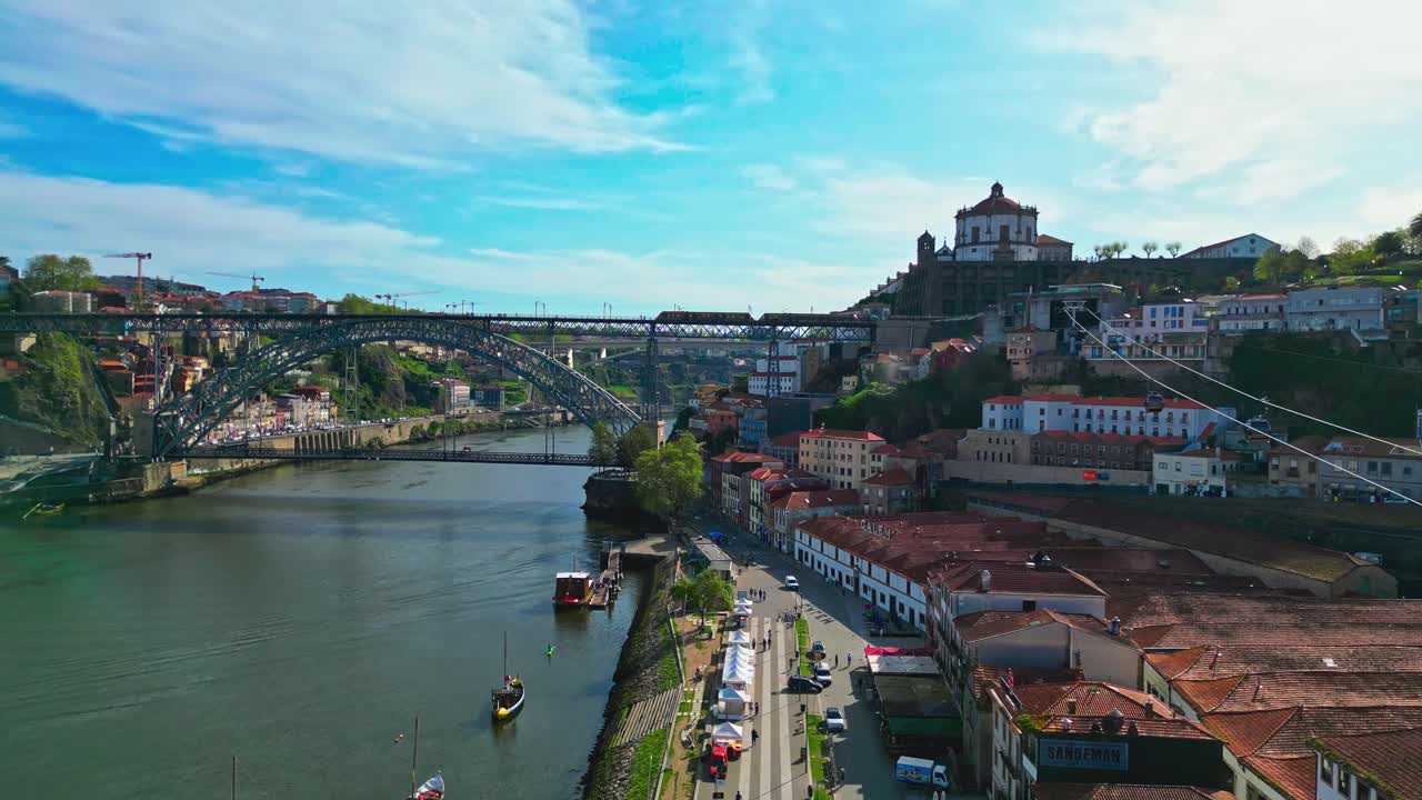 Plano aéreo de acompanhamento do teleférico de Gaia movendo-se ao longo do Rio Douro em direção à Sé do Porto, exibindo a margem do rio e a paisagem urbana histórica.