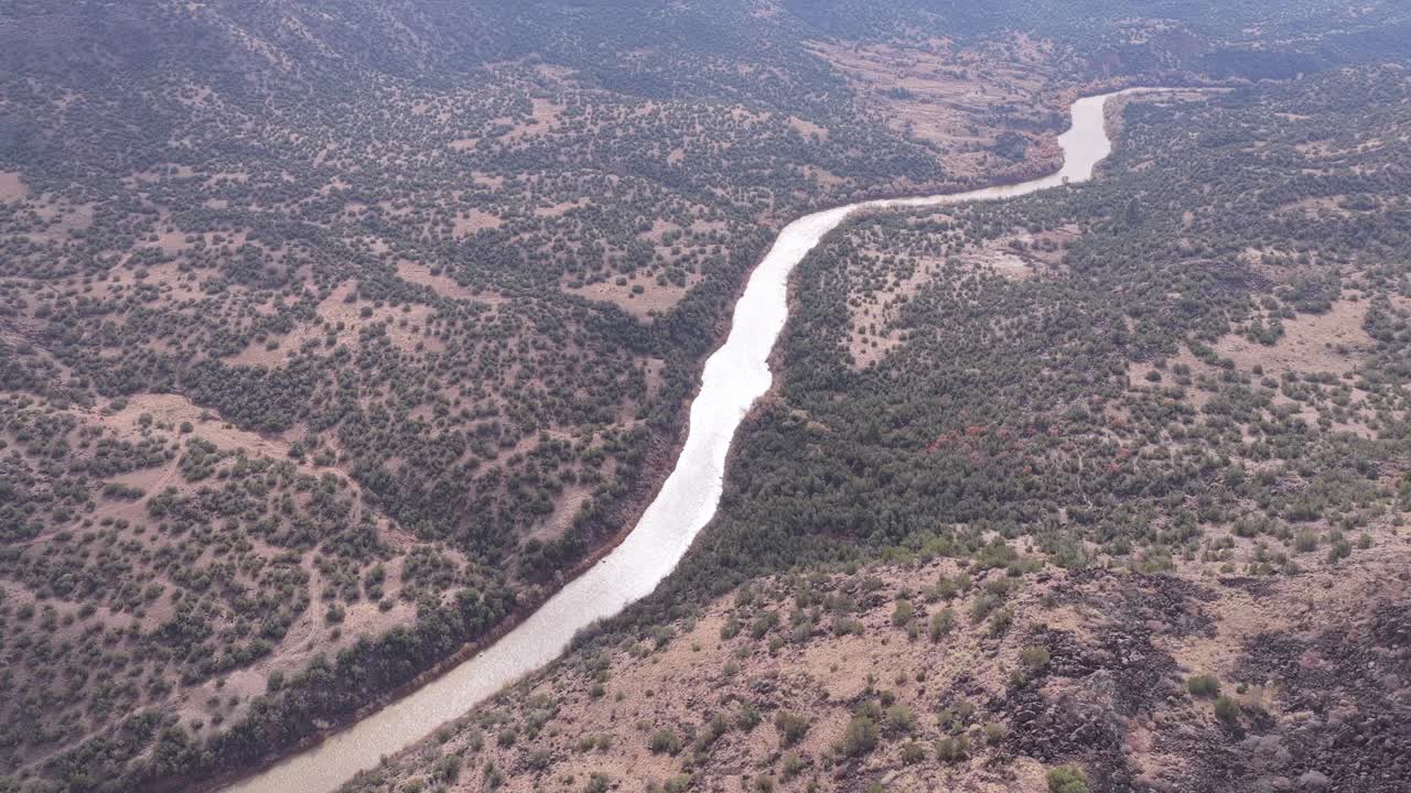High aerial view of the Rio Grande curving through a broad desert canyon with scattered shrubs, rocky slopes, and layered terrain