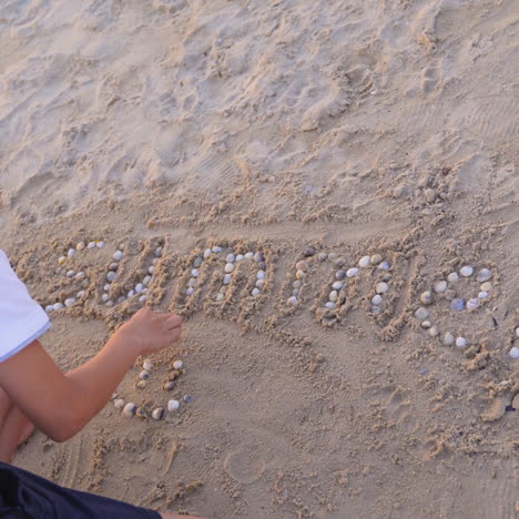 word summer on a sand beach. boy finishes laying out the word summer with small pebbles on sand outdoors.