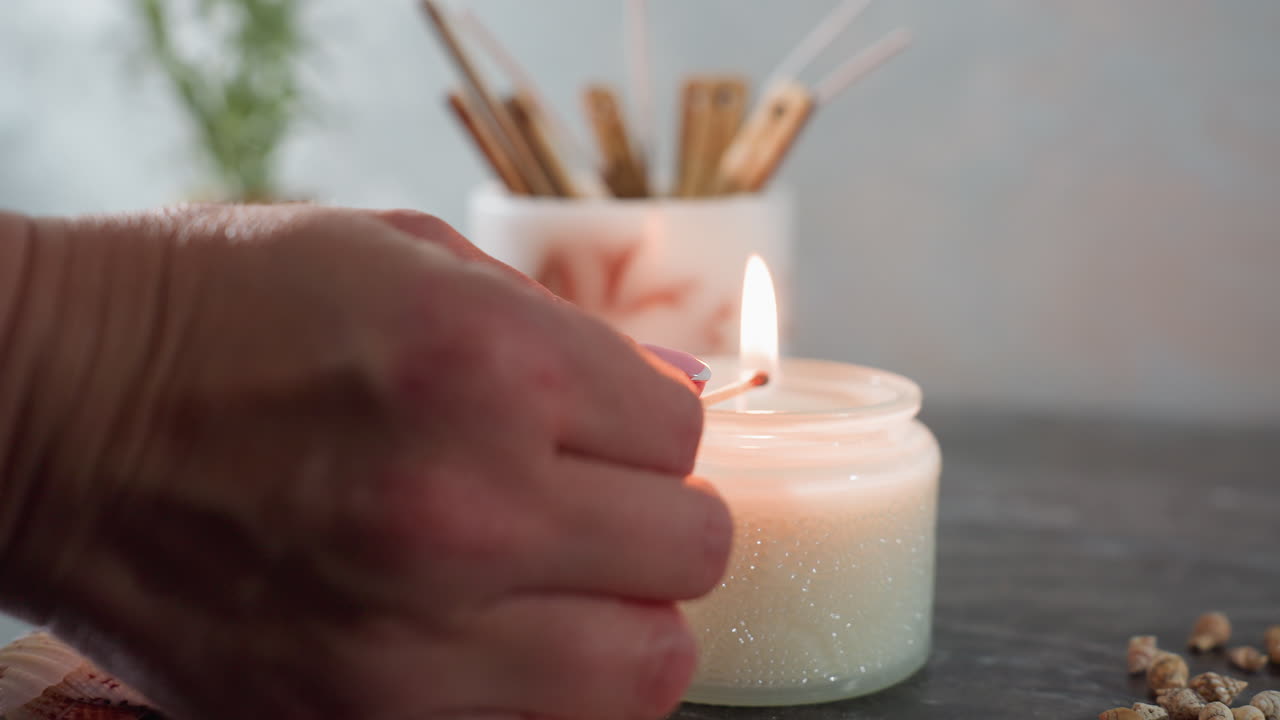 Close up of burning matchstick held by hand approaching white candle surrounded by seashells and snail shells on marble surface with soft background