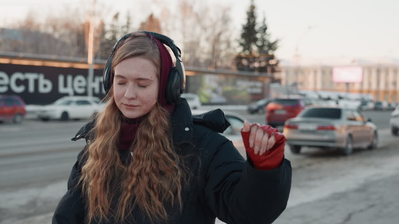 Young white woman headphones snowy roadway adjusting earcup, red gloves and hood up, city traffic and parking lanes behind, introspective morning walk with soft light and chilly air, commuter routine