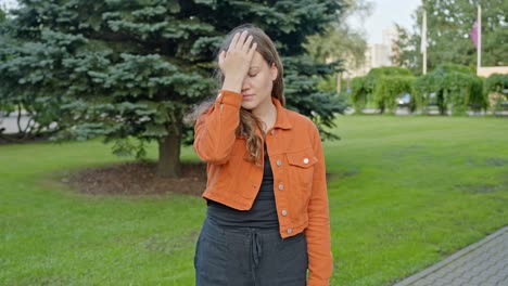 mujer joven haciendo cara de palma en un parque de la ciudad con una emoción olvidadiza vistiendo una chaqueta naranja
