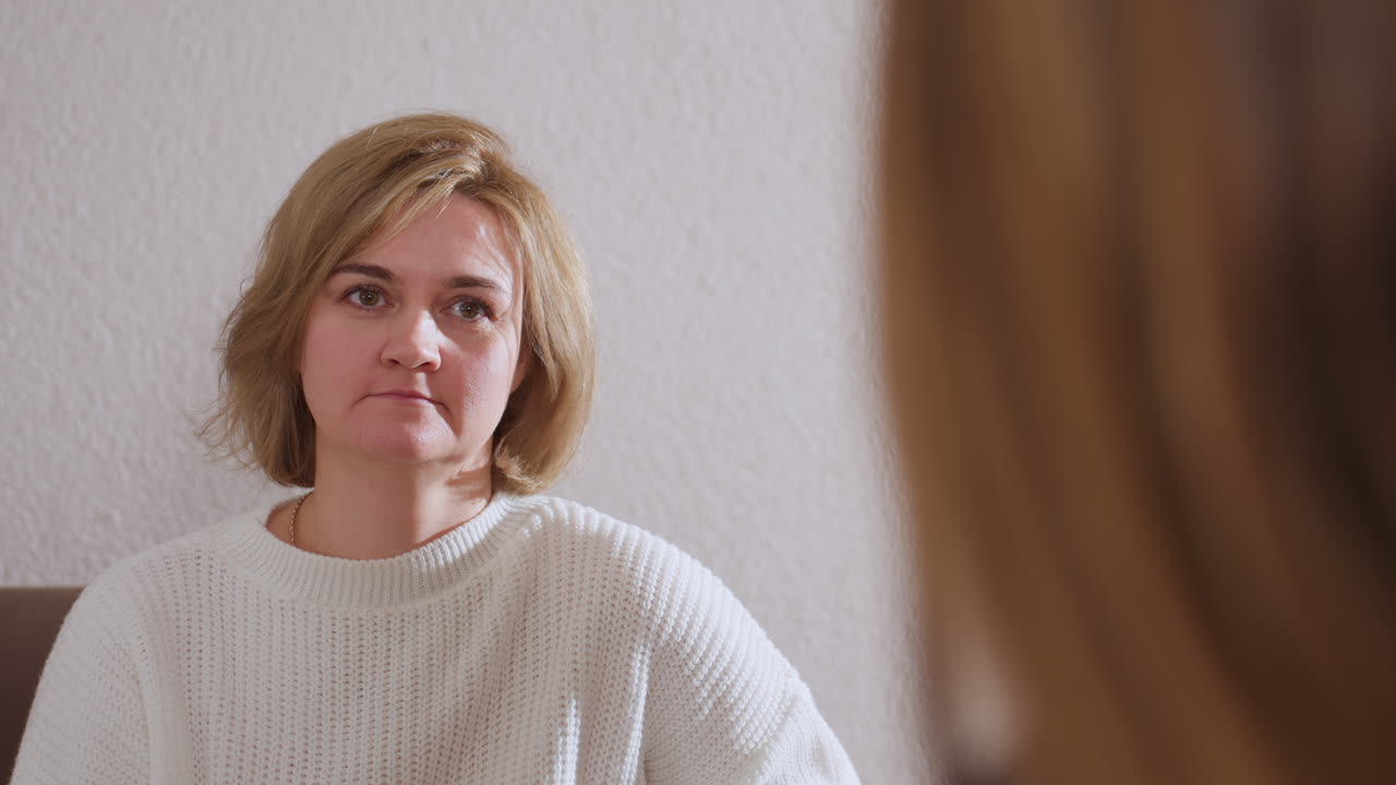 Over shoulder of companion in healing space while guest in white sweater listens attentively with subtle relief on her face, showing empathy, connection, and reflective communication