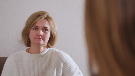 over shoulder of companion in healing space while guest in white sweater listens attentively with subtle relief on her face, showing empathy, connection, and reflective communication