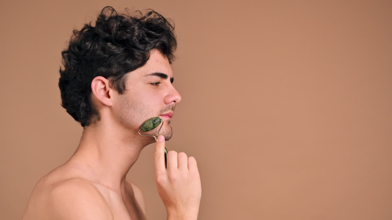 A young caucasian man with stubble beard is doing a facial massage using a roller