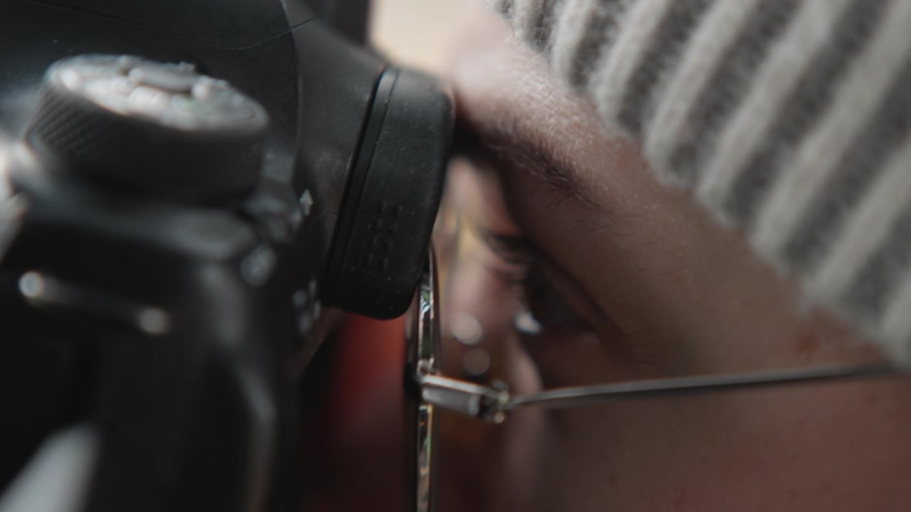 Gros plan à hauteur des yeux, mise au point du viseur du photographe, regard intense à travers l'oculaire, monture de lunettes métallique visible, peau texturée, lumière froide, cadrage précis, concentration technique, professionnel