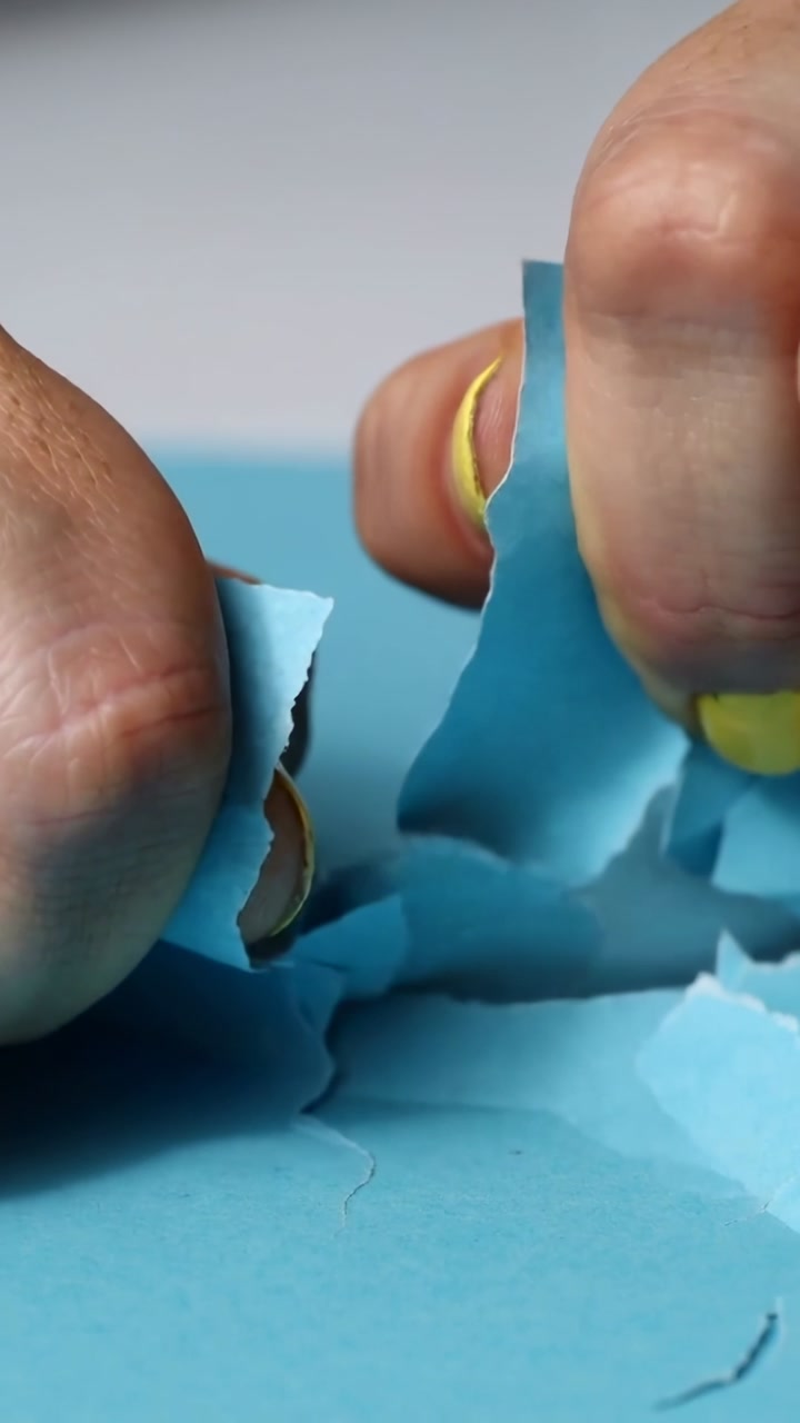 Vertical video: Pinching hands tearing bright blue paper on desk with yellow nail polish and bits