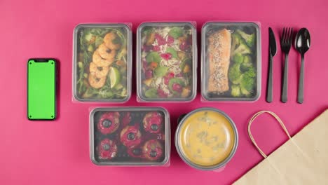 Food delivery top view, take away meals in disposable containers on pink background. Lunch boxes with cooked vegetarian dishes, using phone with chroma green screen. Healthy diet. Catering service