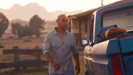 Portrait-Of-Man-Unloading-Backpacks-From-Pick-Up-Truck-On-Road-Trip-To-Cabin-In-Countryside