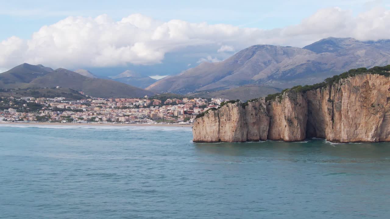 Ocean view of seaside Gaeta town and Montagna Spaccata outcropping with mountains in background on sunny day, Italy, above aerial approach