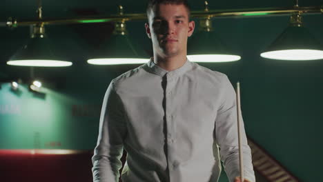 el jugador de billar con camisa blanca se sienta en una posición enfocada, sosteniendo el palo de la clave antes de levantarse con cara seria. la sala de billar débilmente iluminada mejora la concentración, la estrategia y la atmósfera competitiva