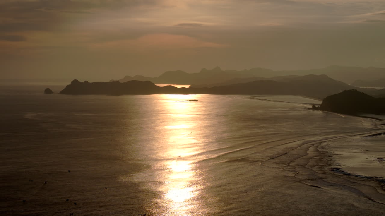 황금빛의 해가 지는 곳 은 조용 한 해안 해역 에 반사 되어 있으며, 배경 에는 멀리 있는 산 들 이 있다