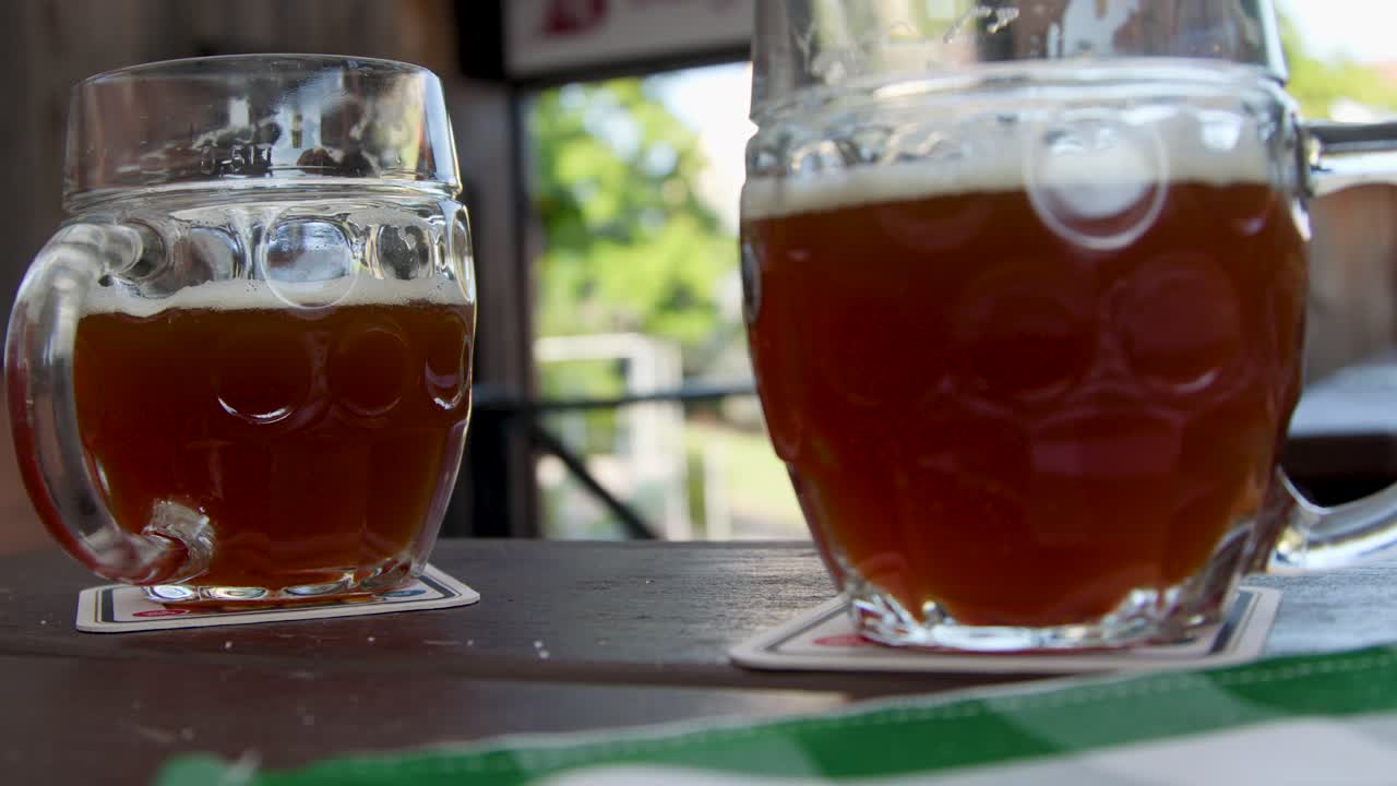 Hand lifts traditional beer mug from wooden table in sunlit outdoor beer garden, natural daylight