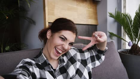 chica morena feliz vestida con camisa a cuadros sonriendo, posando, hace señales de paz con los dedos haciendo autorretrato - punto de vista de la cámara. cámara lenta
