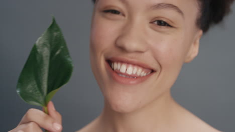 Primer-Plano-Retrato-Hermosa-Mujer-De-Raza-Mixta-Sonriendo-Acariciando-La-Cara-Con-Hojas-Tocando-Una-Tez-De-Piel-Suave-Y-Saludable-Disfrutando-De-La-Belleza-Natural-Del-Cuidado-De-La-Piel-Sobre-Fondo-Gris