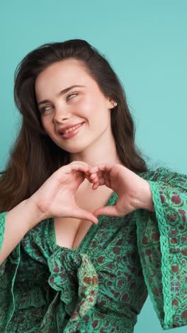 happy woman blowing kiss and showing heart gesture in blue studio