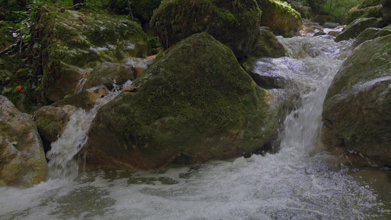 Nærbillede i slow motion af et sprøjtende lille vandfald i Schweiz' vilde natur - 4k panorering i grønne bjerge med mos og træer om sommeren