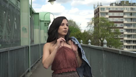 slow motion of an attractive and playful latina woman with black wavy hair and a jean jacket walking on a bridge in london, looking at the camera, happy with a beautiful smile