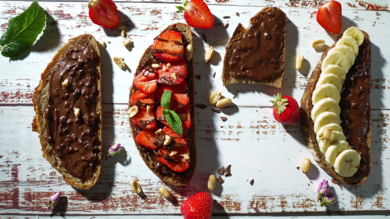 A hand places and holds a bitten slice of chocolate spread toast, surrounded by full slices topped with strawberries, bananas, and peanuts, on a rustic white wooden table.