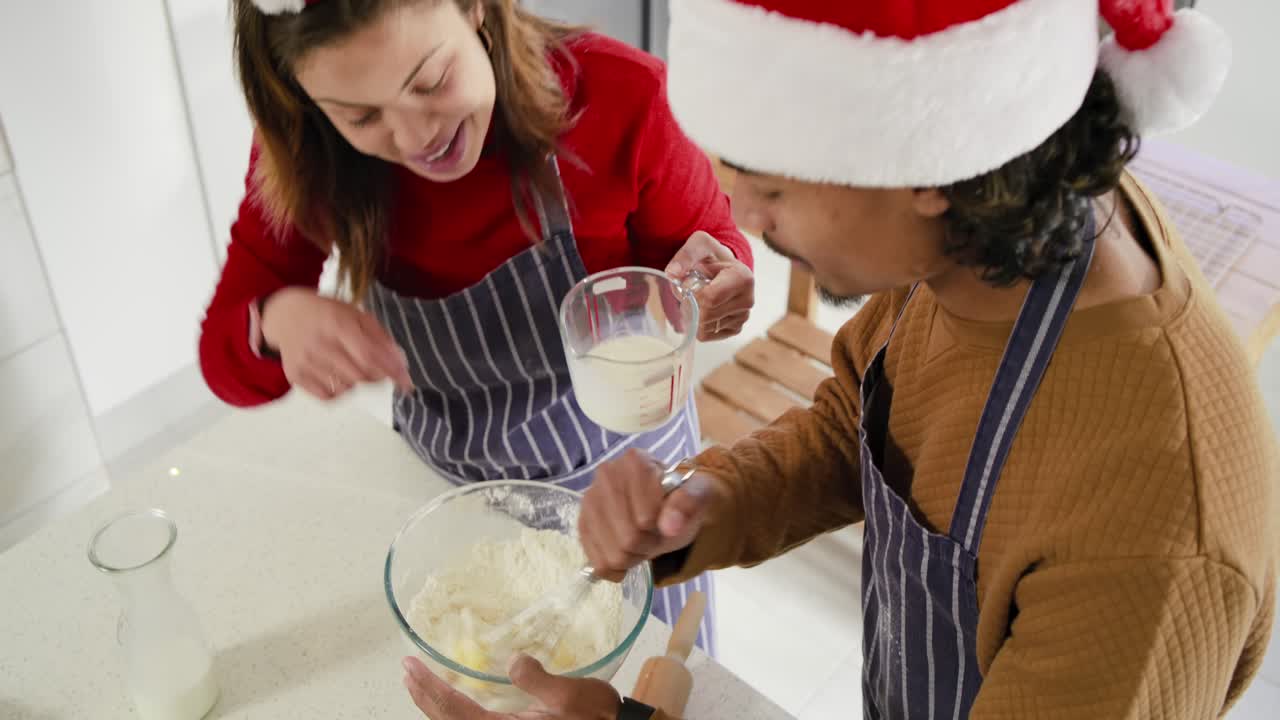 Couple wearing Santa hat whisking batter in home kitchen after lifting cup for holiday baking