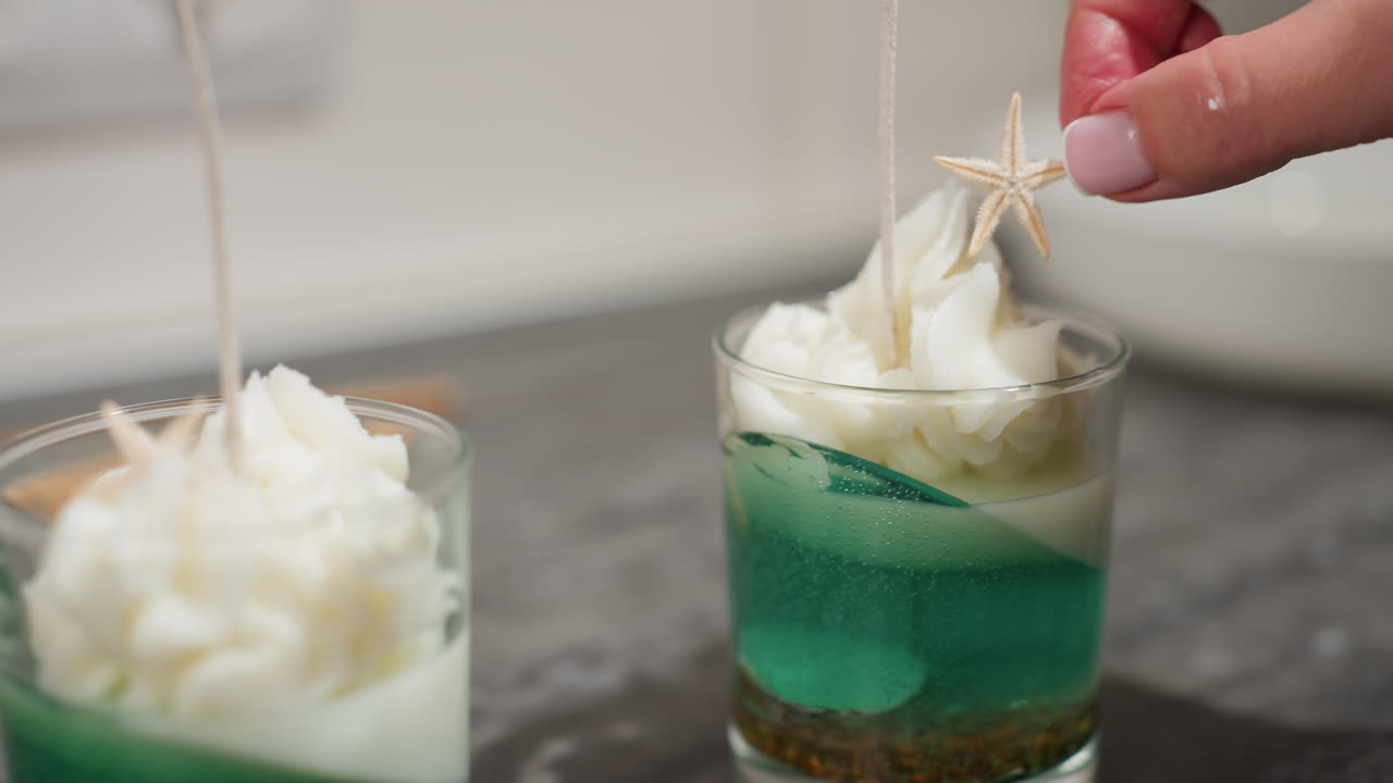 Pastry chef carefully places star decoration atop ice cream swirl in transparent dessert cup filled with green and white layered treat, showcasing intricate dessert presentation with hand gesture