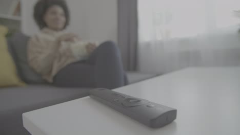 detail of a remote control. in the background, a young african american woman enjoys watching a movie and eats popcorn