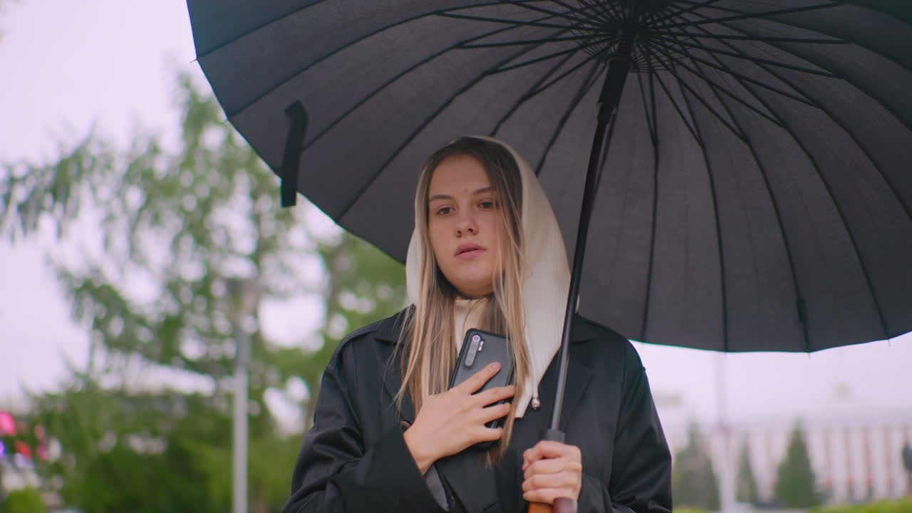 Young woman under umbrella wearing hooded coat talks on phone during rainy day outdoors, looking down with broken expression, surrounded by blurred greenery and city background atmosphere