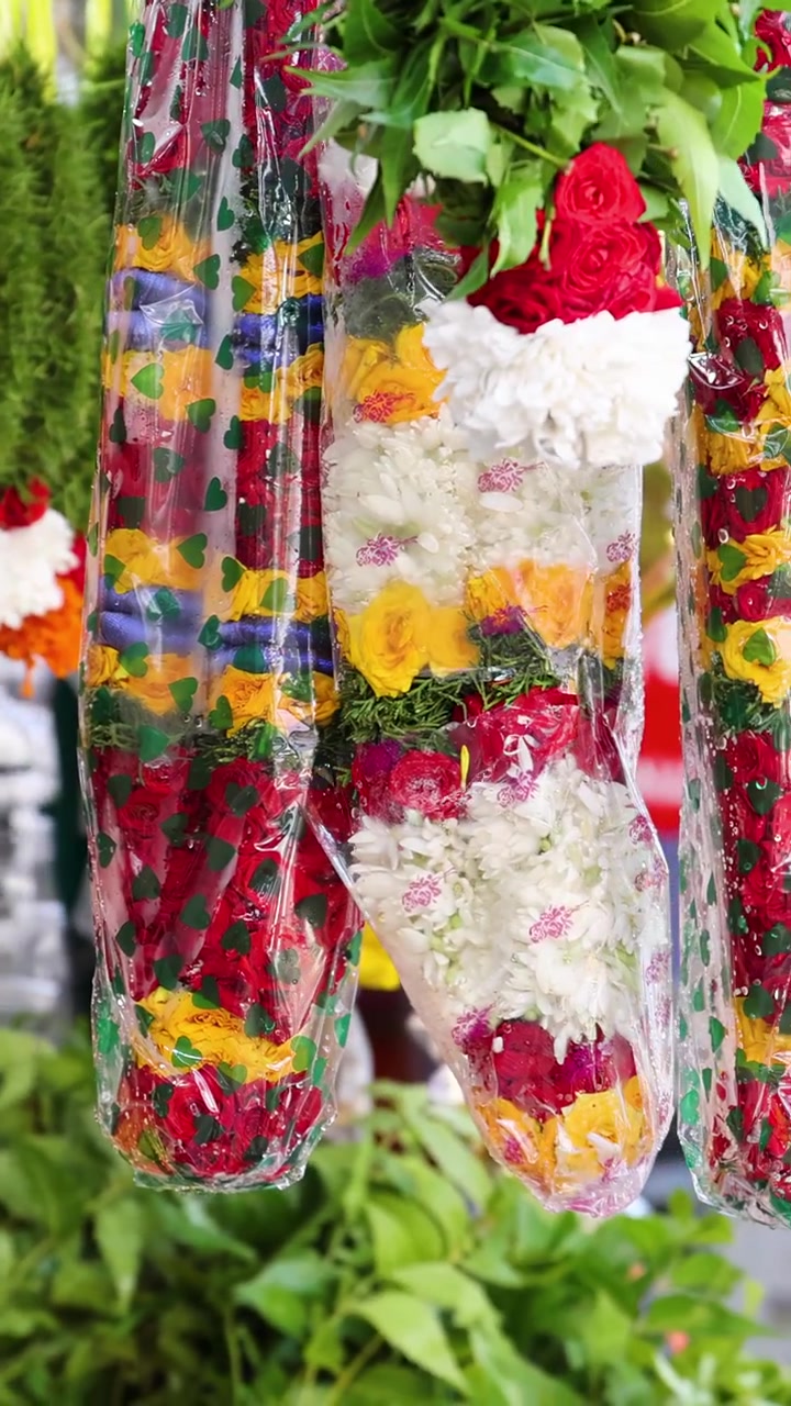Ghirlande di fiori dai colori vivaci, avvolte nella plastica, ruotano lentamente in esposizione su una bancarella di un mercato all'aperto. Luce naturale, profondità di campo ridotta, atmosfera vibrante.