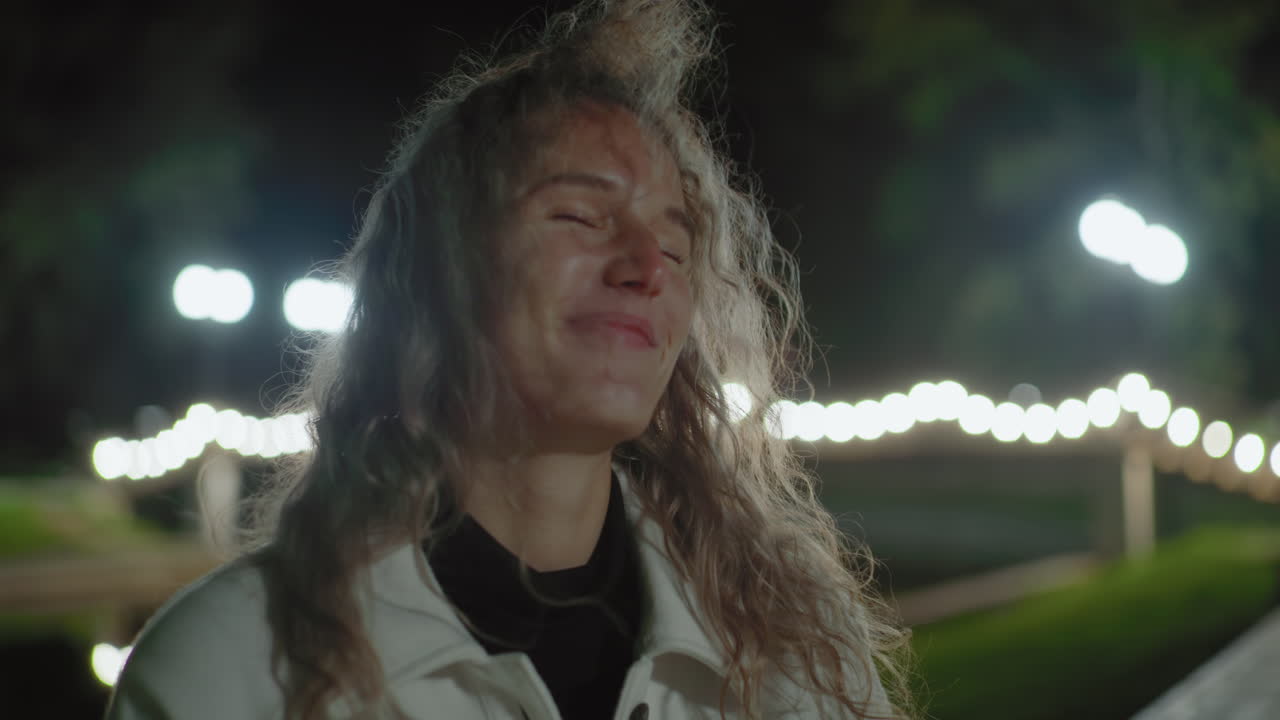 Joyful white lady with curly hair seated outdoors at night, hair swaying gently under warm lights, blurred reflection behind, calm ambiance capturing bright smile in peaceful environment