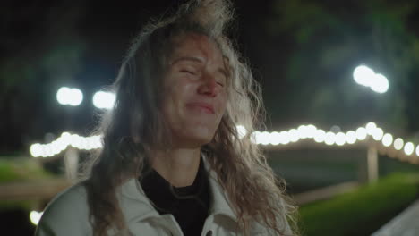 joyful white lady with curly hair seated outdoors at night, hair swaying gently under warm lights, blurred reflection behind, calm ambiance capturing bright smile in peaceful environment