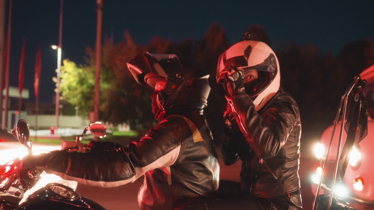 Lady lifts visor smiling and gesturing playfully while seated behind partner on motorcycle, both wearing helmets and leather gear, waiting at traffic stop under bright city lights