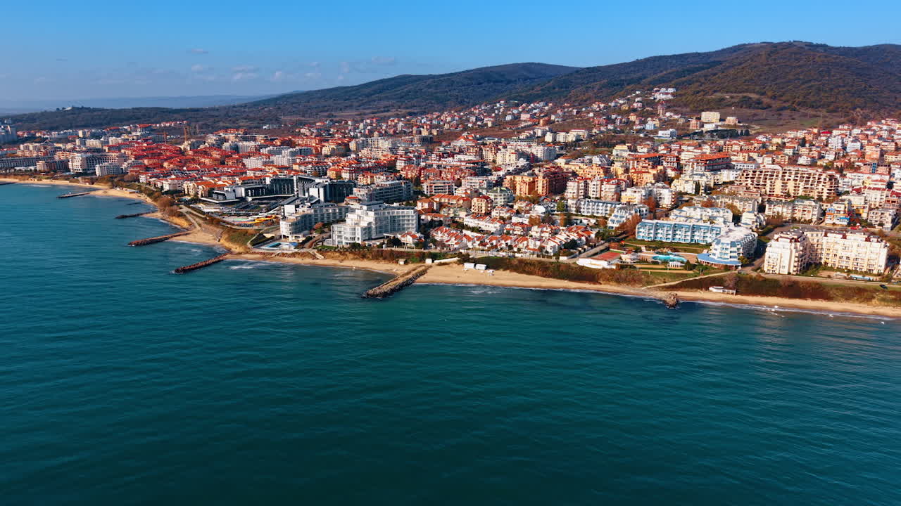 Deniz kenarında, suya sıfır bir sahil kasabası. Binalar kıyı şeridini kaplarken, arka planda tepeler yer alıyor. Güneş, ev ve otel sıralarını aydınlatarak manzaranın üzerine parlıyor.