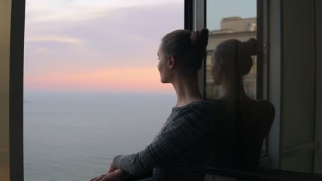 closeup view of beautiful young woman standing by the open window during the sunset by the sea and enjoying the view